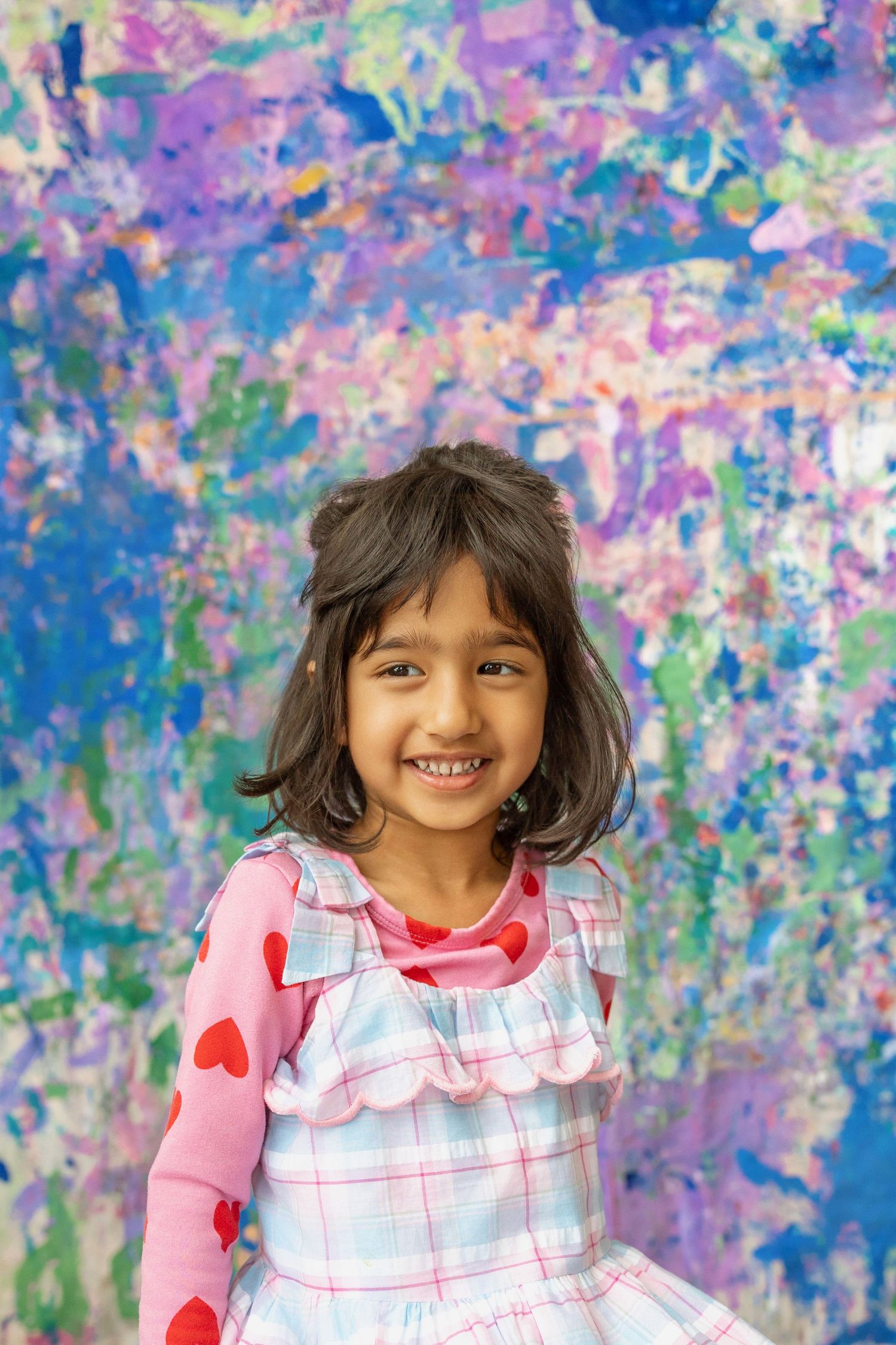 A happy young girl in a plaid dress smiles brightly against a colorful painted backdrop.
