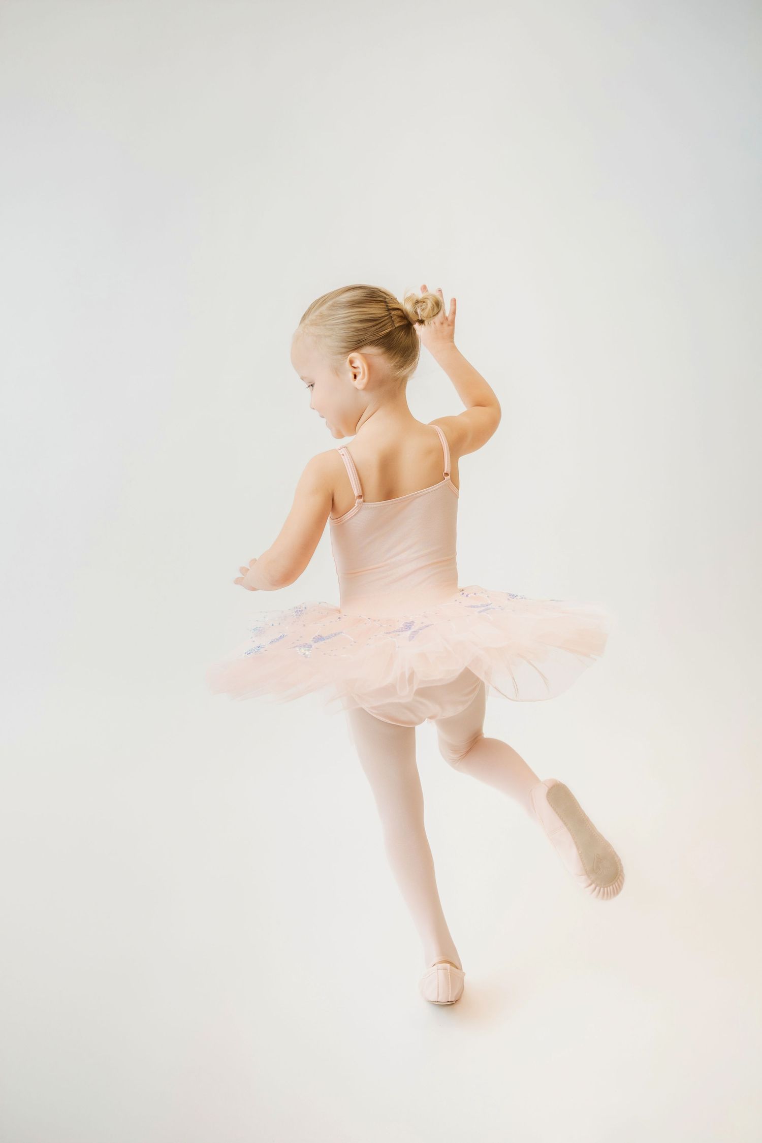 Young ballerina in pink tutu dancing on tiptoe with hand raised to hair, viewed from behind.
