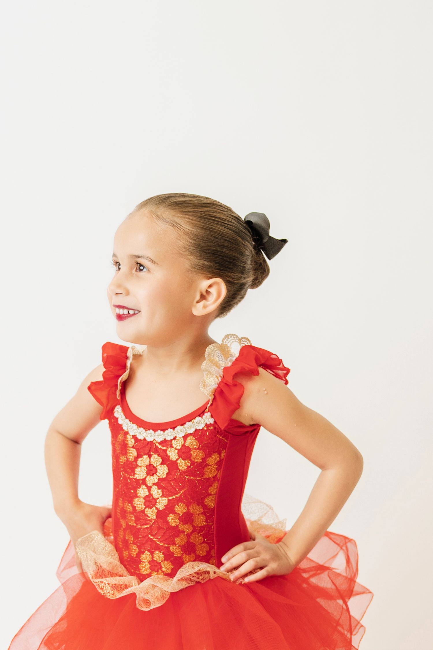 Young ballerina in red and gold floral tutu poses with hands on hips, smiling in a white studio.