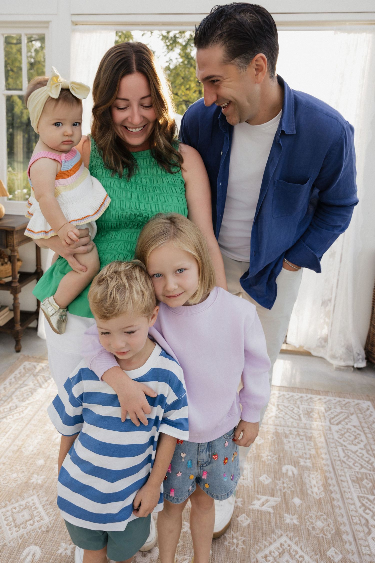 Happy family of five smiling indoors, parents with three young children dressed in colorful casual clothing.