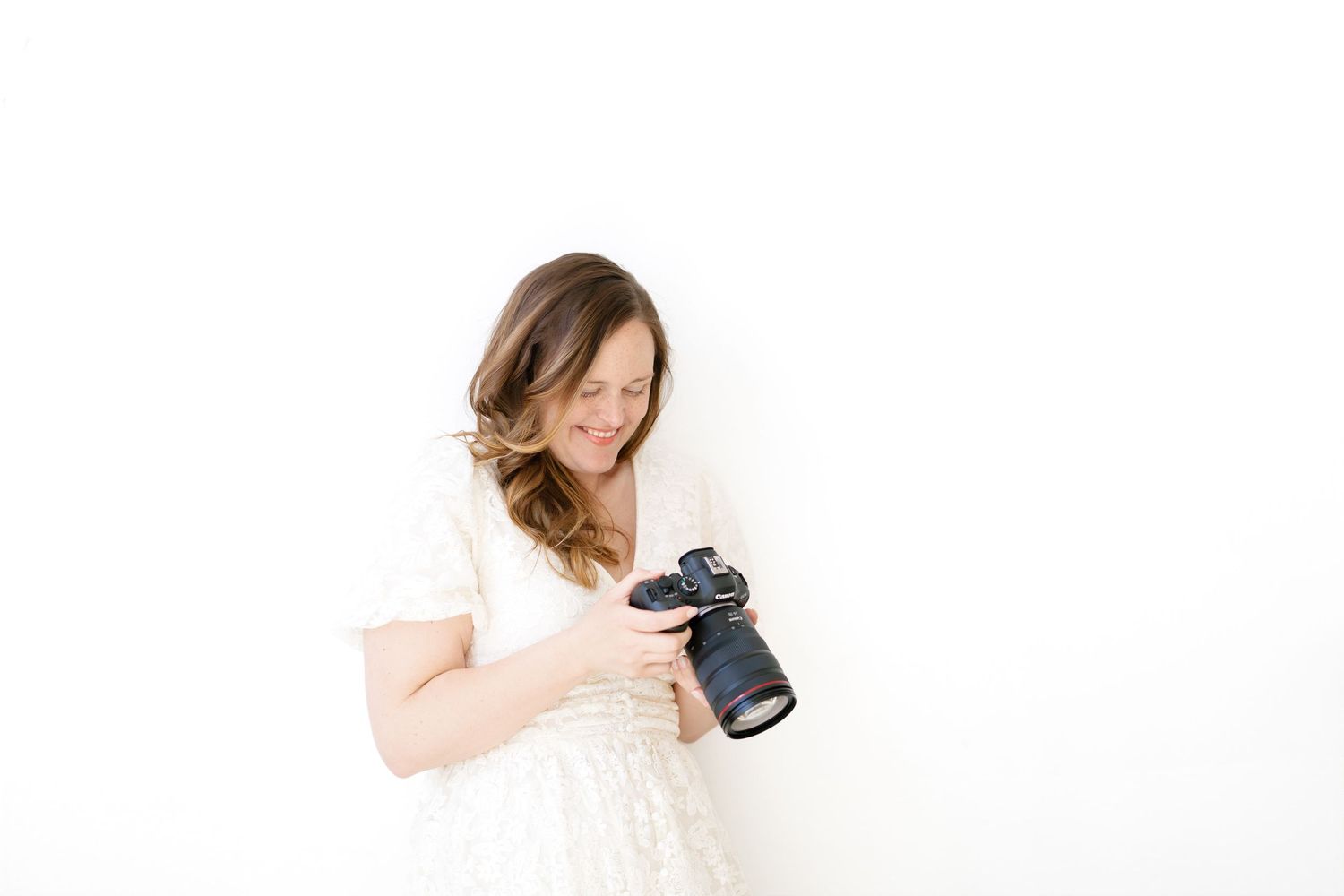 A smiling woman in a white lace dress holds a professional DSLR camera, reviewing photos against a white background.