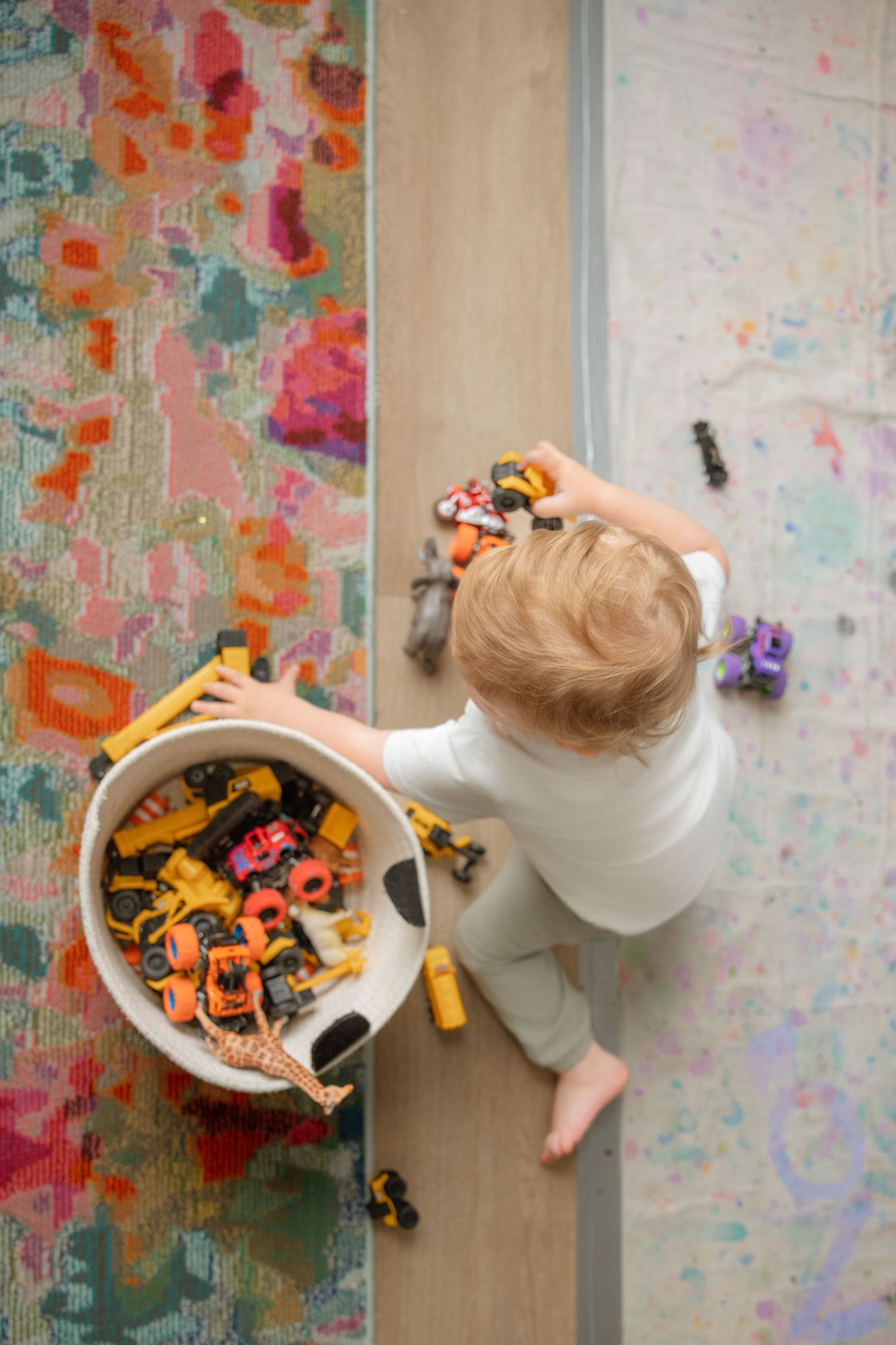 Overhead view of a toddler playing with colorful toy vehicles on a hardwood floor beside a vibrant area rug.