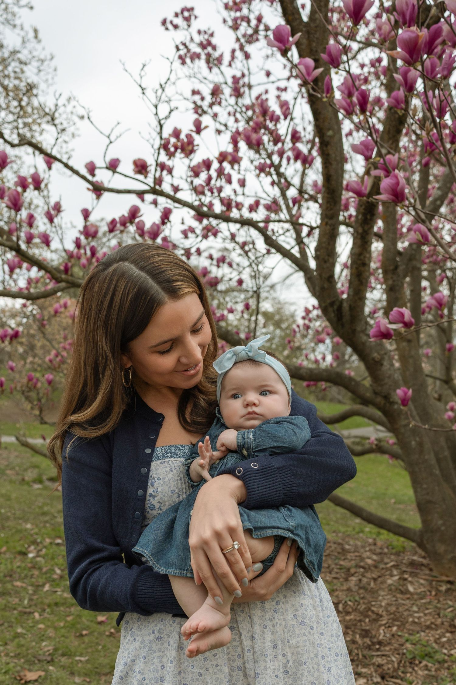 Mother tenderly holding newborn baby near blooming pink magnolia tree.