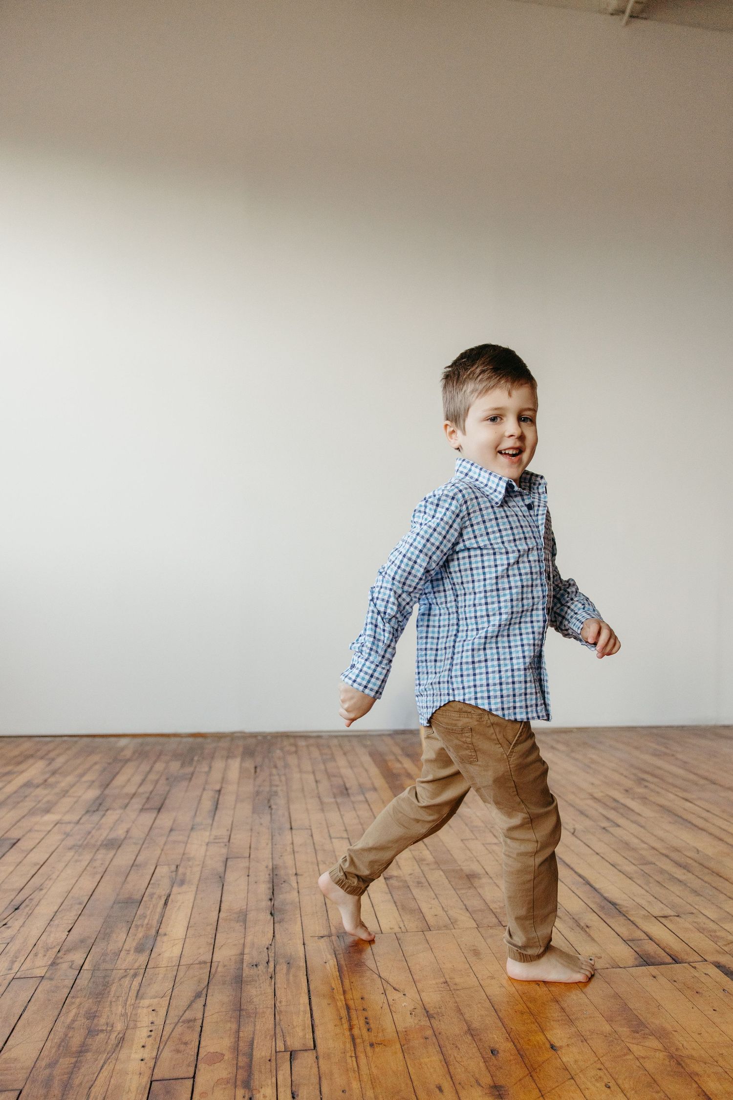 Happy barefoot boy in a plaid shirt and tan pants runs and smiles in a bright, open wooden-floored room.