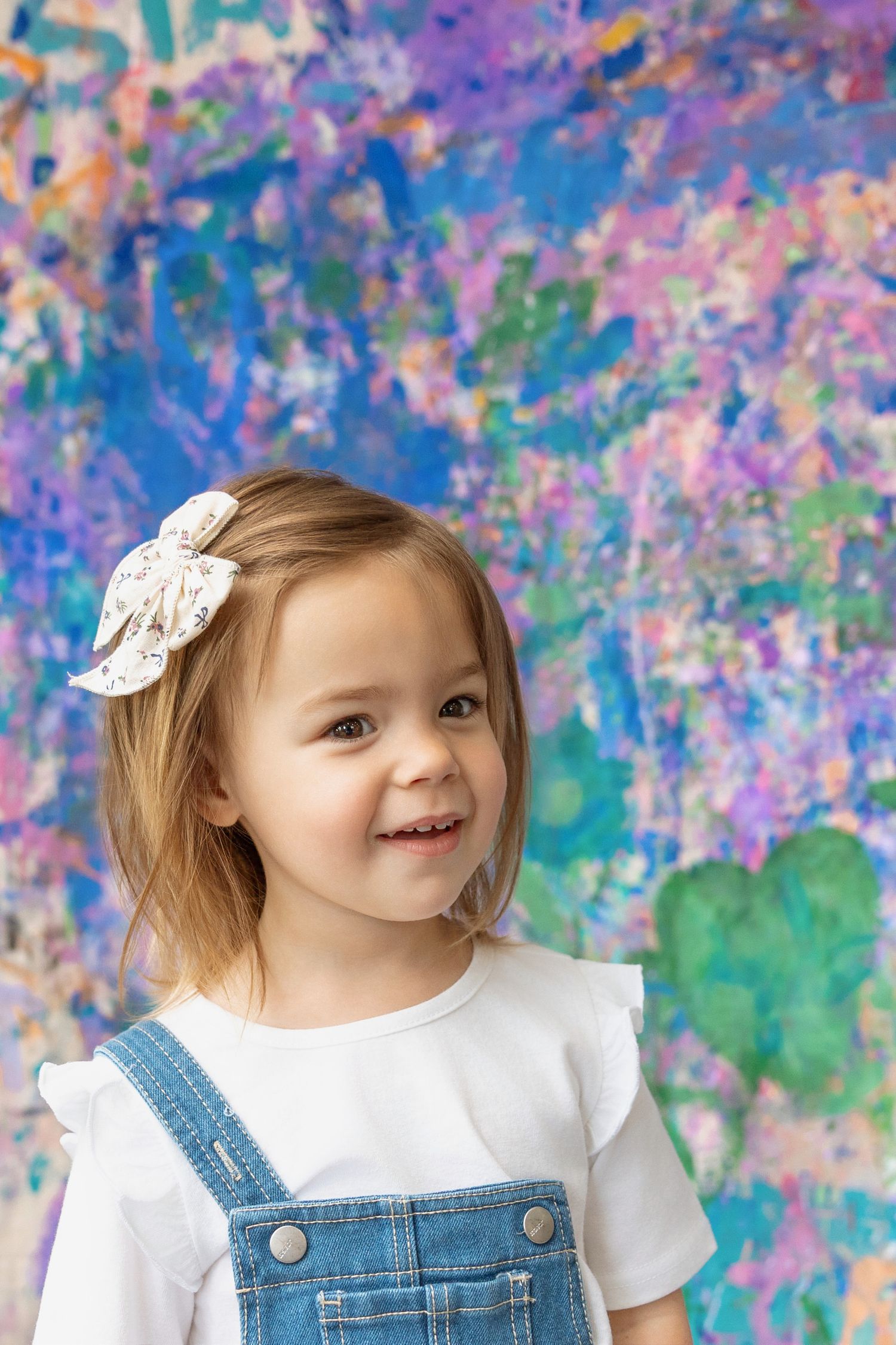 A cheerful toddler girl with a bow in her hair smiles against a colorful painted background.