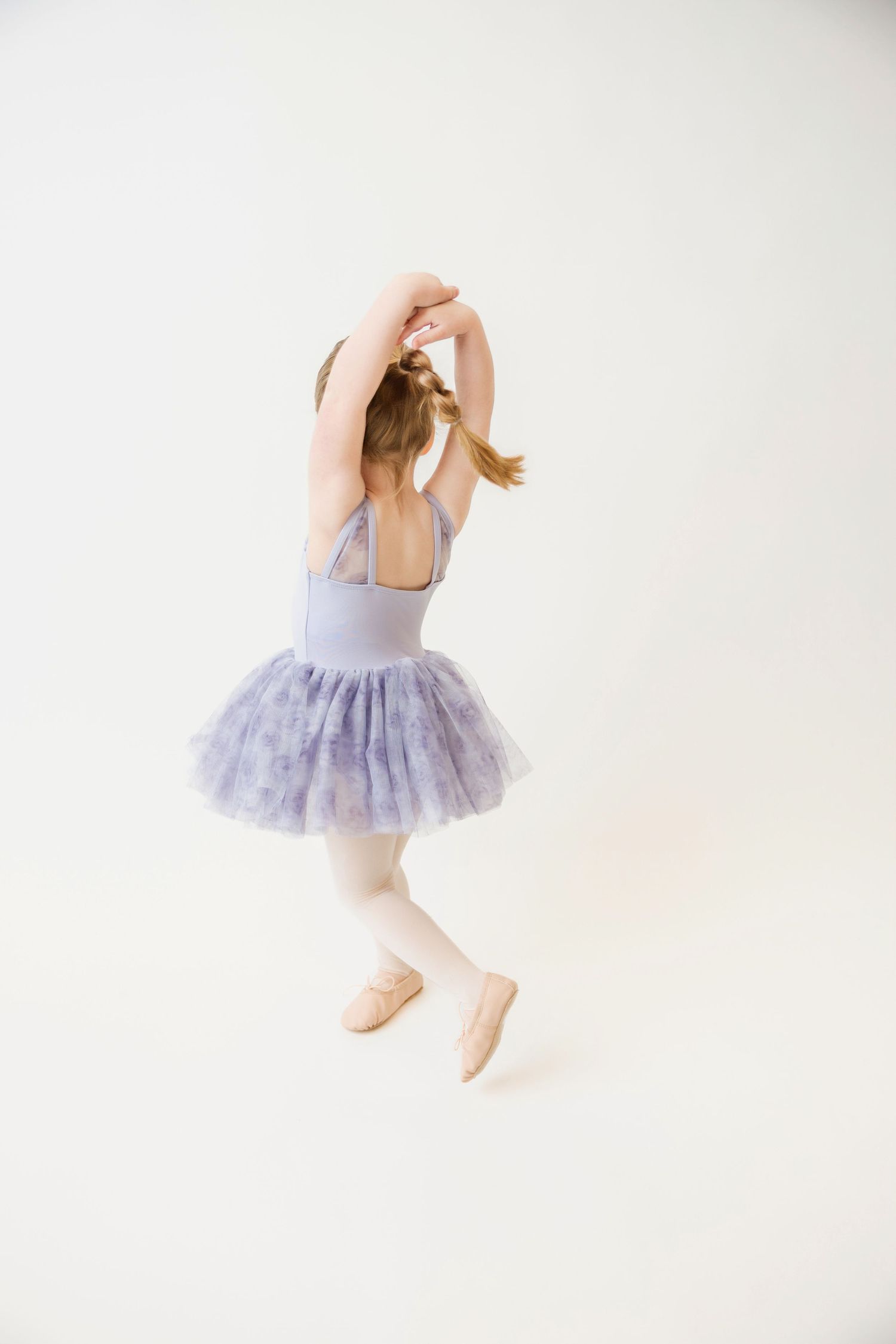 Young ballerina in purple floral tutu dancing with arms raised overhead, photographed from behind.