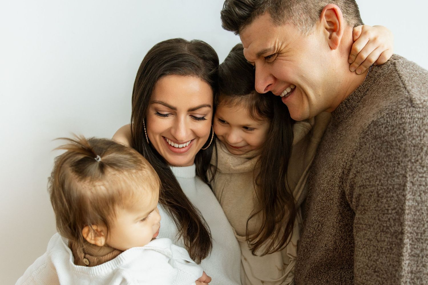 A happy family of four, two parents and two daughters, share a warm, joyful embrace against a light background.