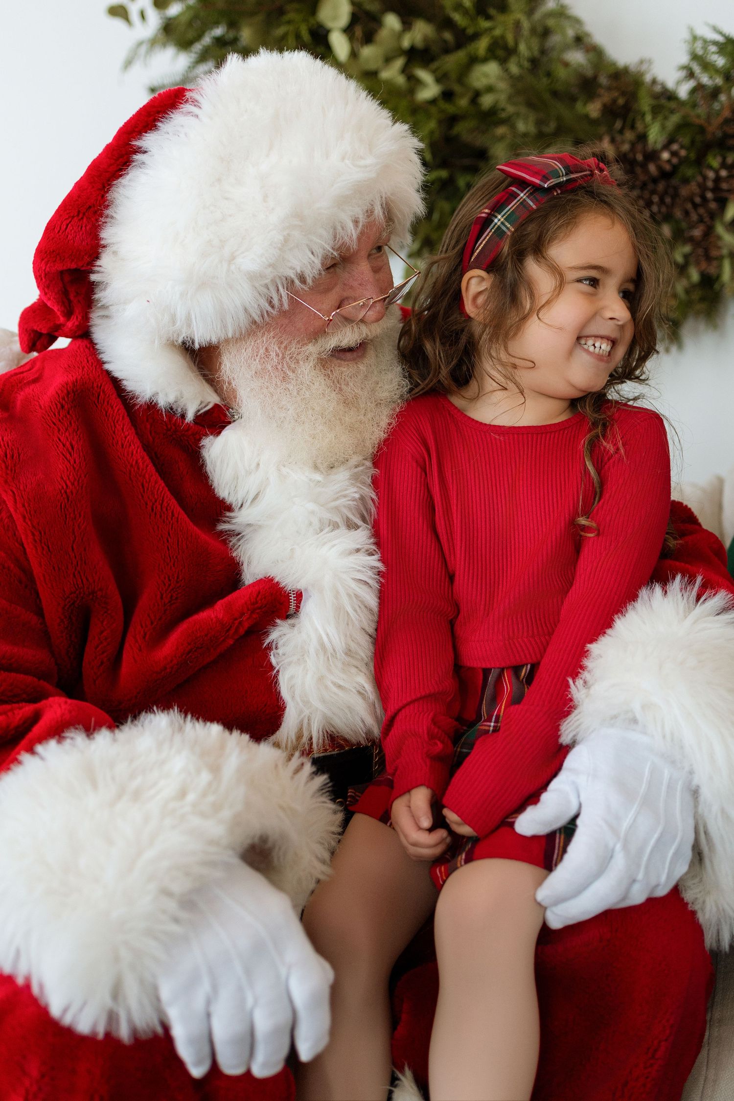 Joyful young girl in red outfit laughs while sitting on Santa Clauss lap during Christmas photos.