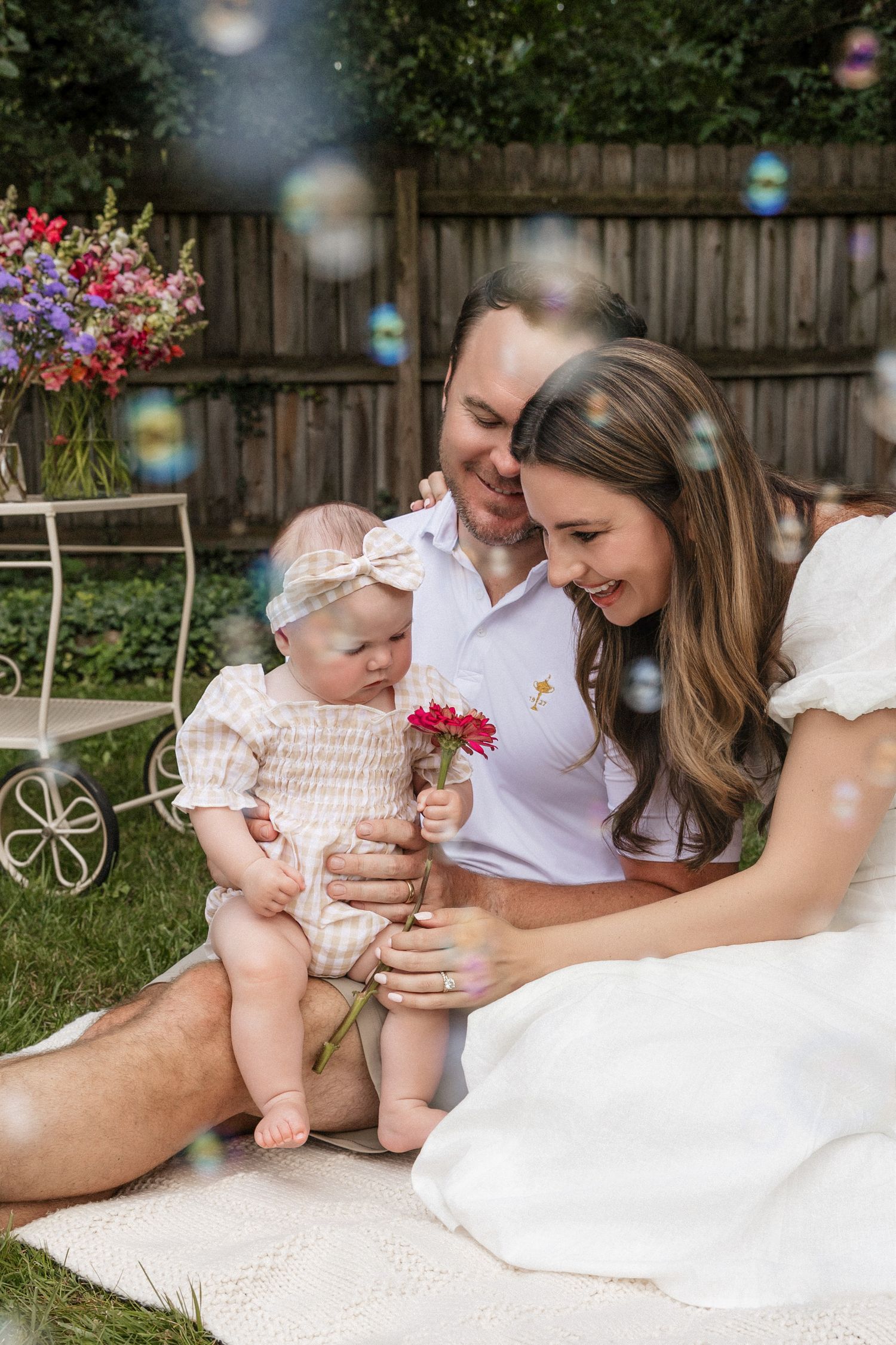Happy young family with baby girl sitting outdoors in garden surrounded by soap bubbles and colorful flowers.