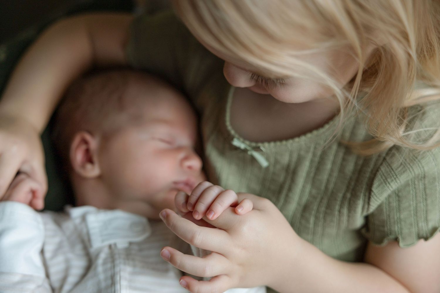 Blonde toddler girl gently holds sleeping newborn baby's hand in a tender sibling bonding moment.