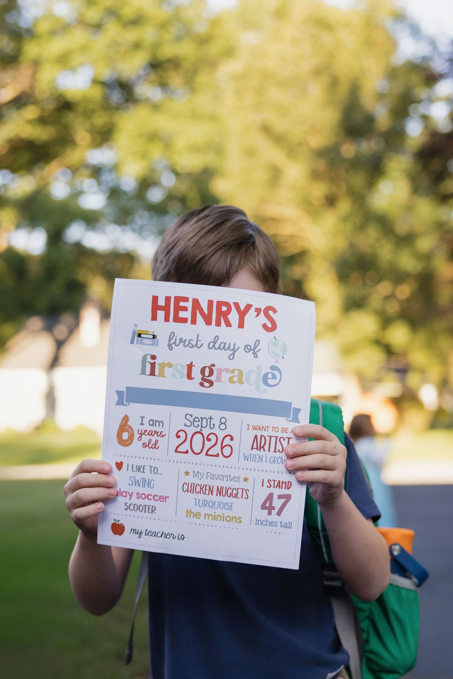 A young boy holds up a colorful first day of first grade sign with his name Henry, wearing a backpack outdoors.
