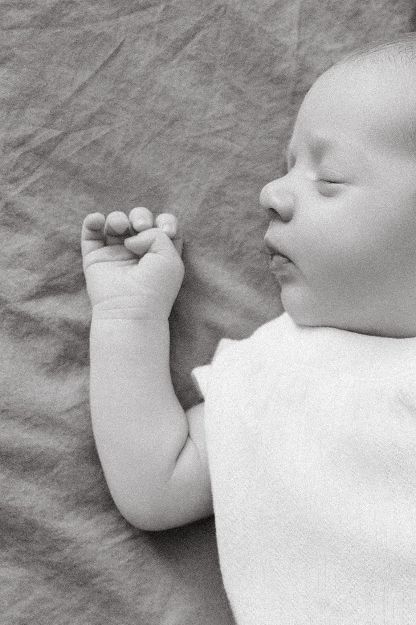 A peaceful sleeping newborn baby in white onesie with tiny fist raised in black and white photo.