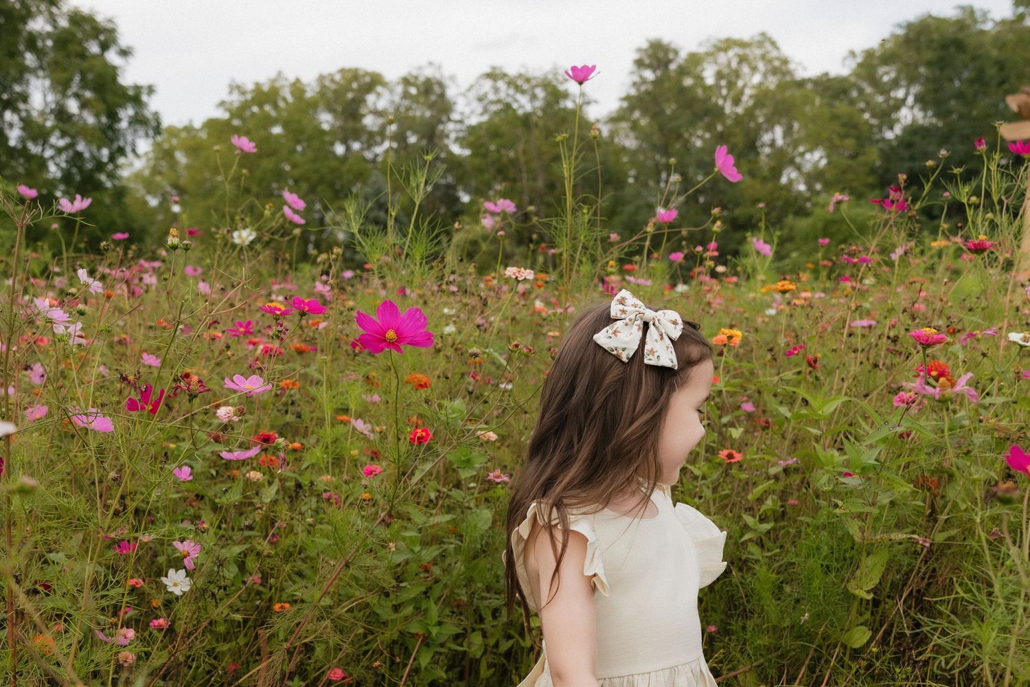 A young girl with a bow in her hair stands among vibrant pink wildflowers in a lush garden field.