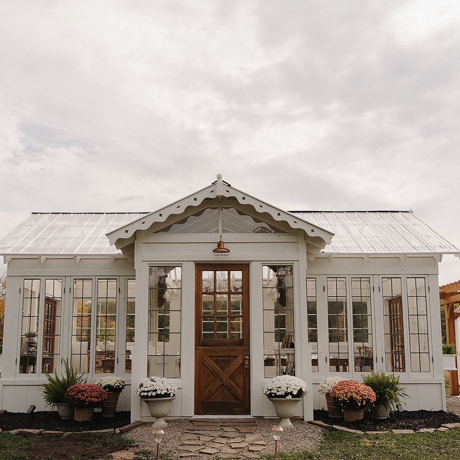Charming white greenhouse with wood Dutch door, arched windows, and flower pots under overcast sky.
