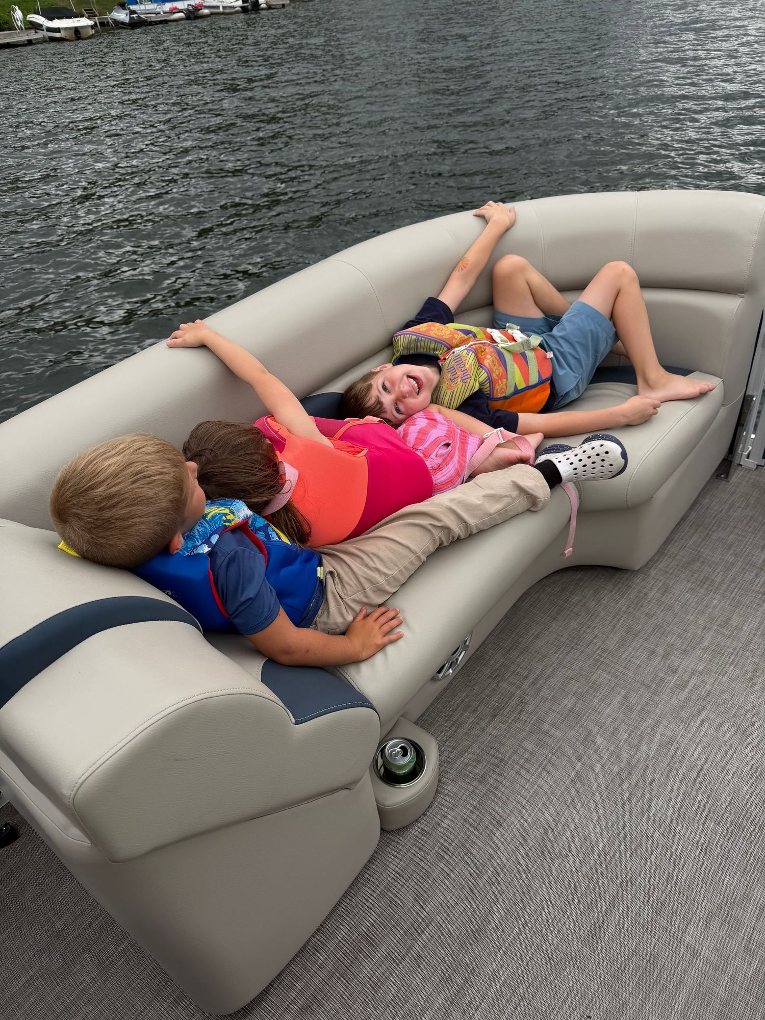 Three children wearing life jackets relax on a pontoon boat couch near the water on a sunny day.