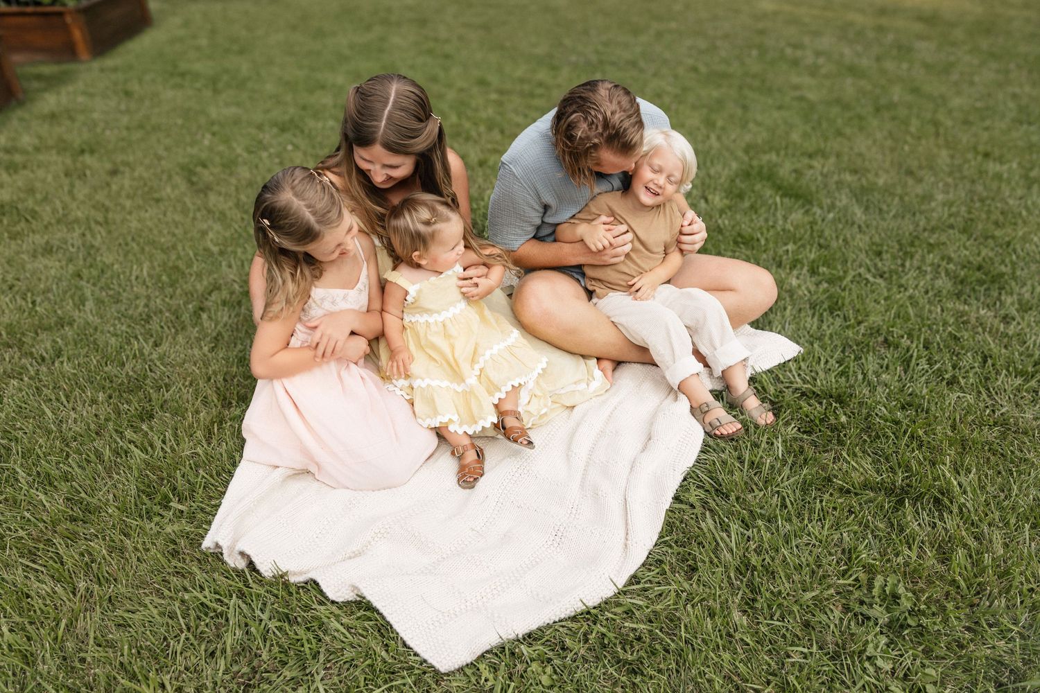 Family of five sitting on white blanket in green grass, parents and three young daughters in pastel dresses sharing a joyful moment.