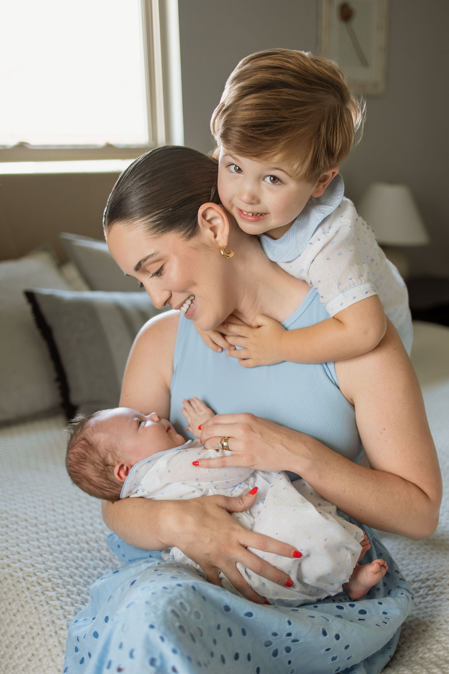 Happy mother sitting on bed holding newborn while older toddler child hugs her from behind.