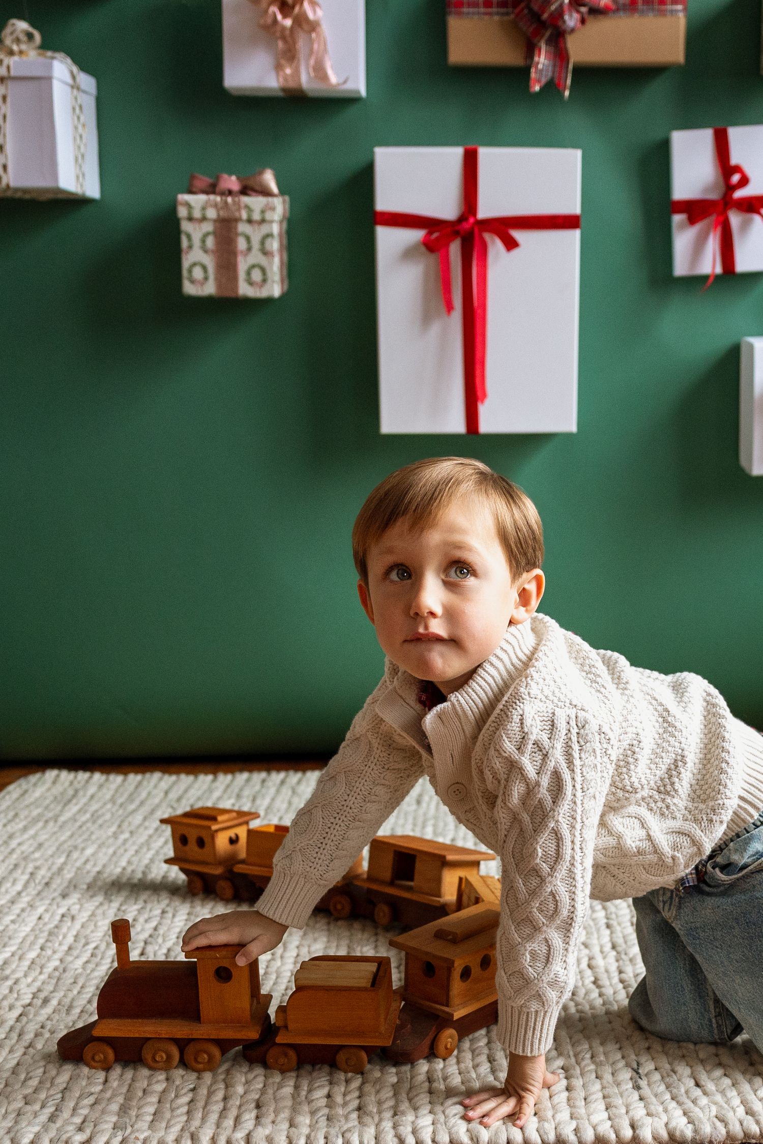 Young boy in cream knit sweater playing with wooden toy train set at Christmas.