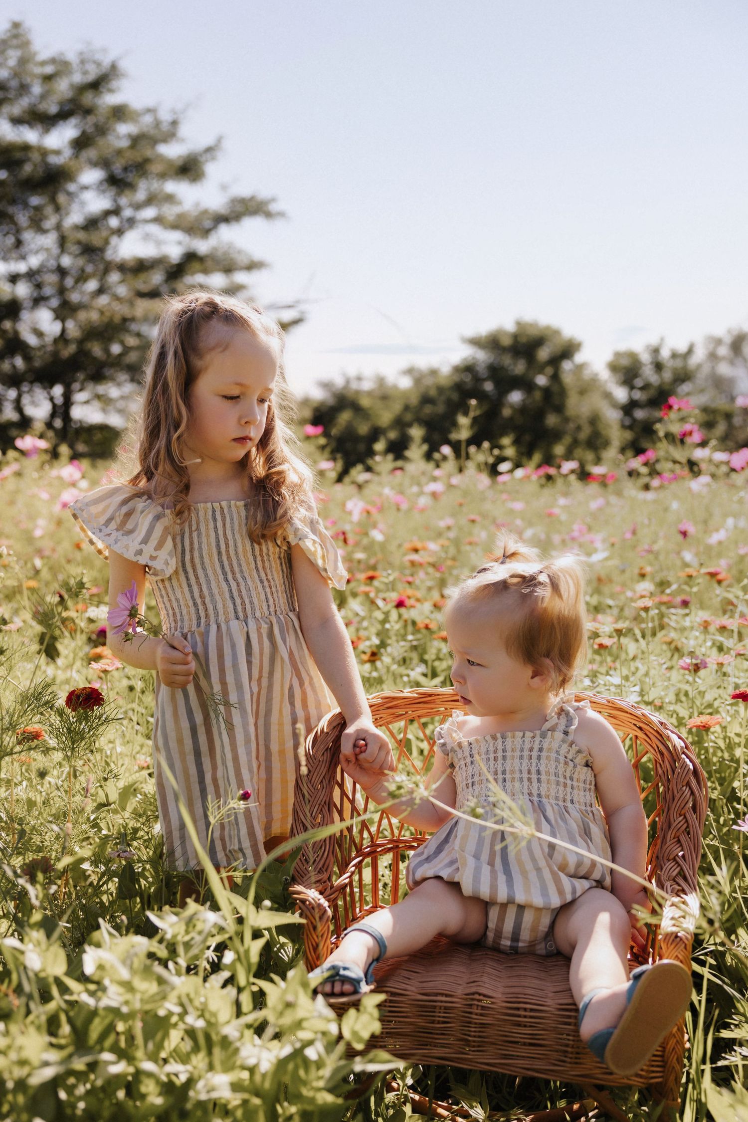 Two young girls in striped dresses play in a colorful flower field on a sunny day.