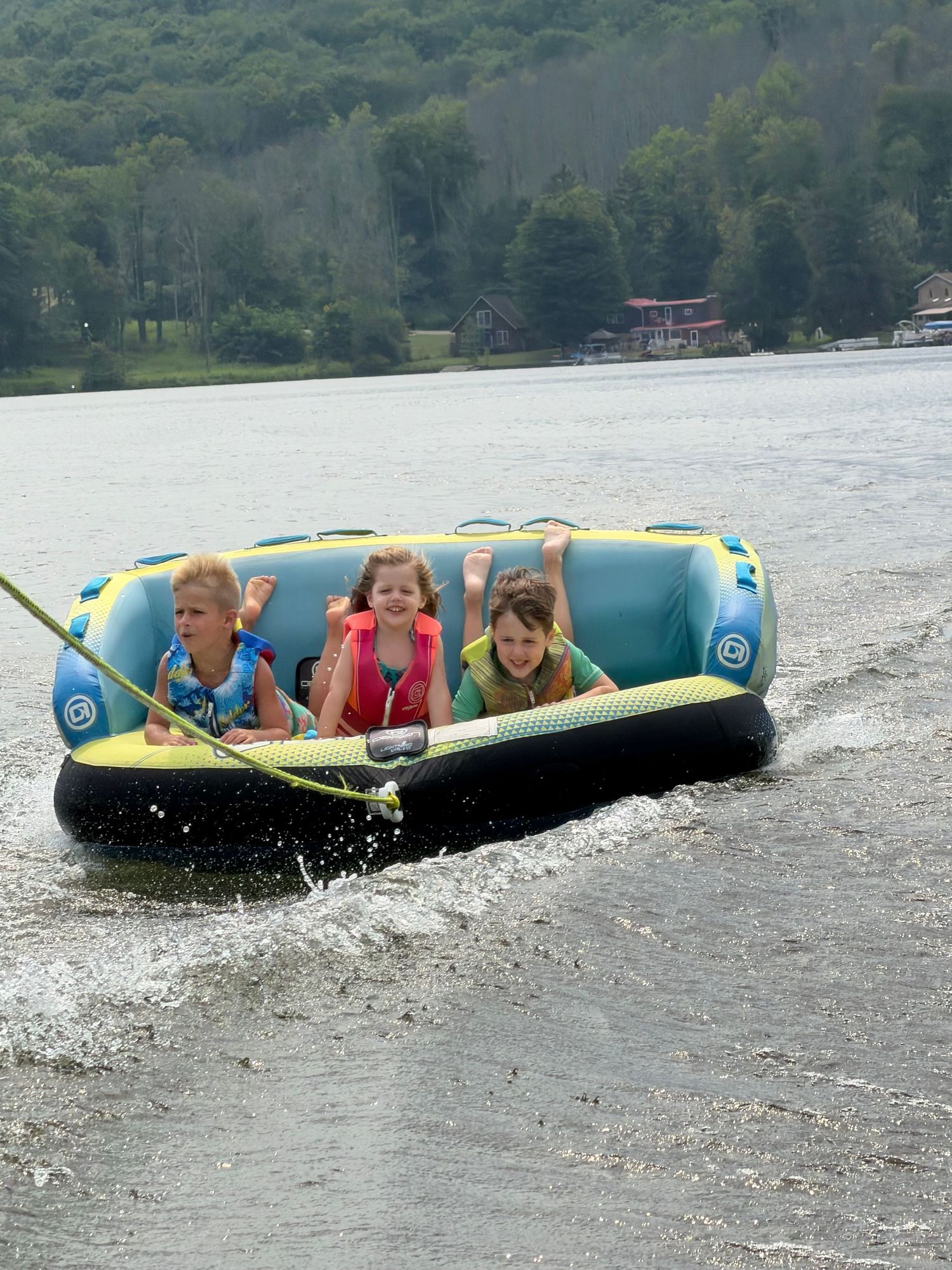 Three children riding a towable tube on a lake, wearing life jackets, smiling and waving while being pulled by a boat.