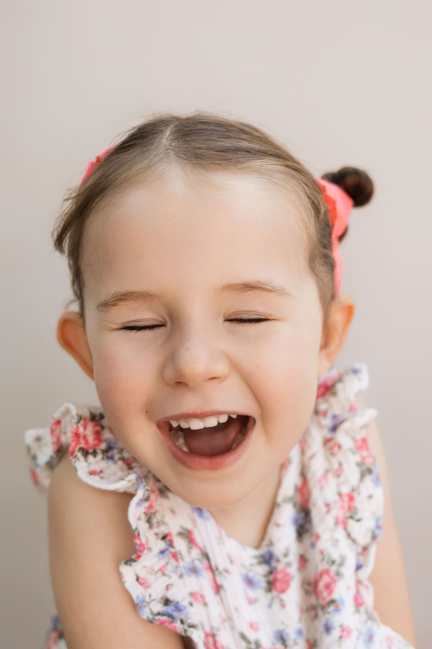 Joyful toddler girl laughing with eyes closed, wearing a floral dress with pink hair ties.