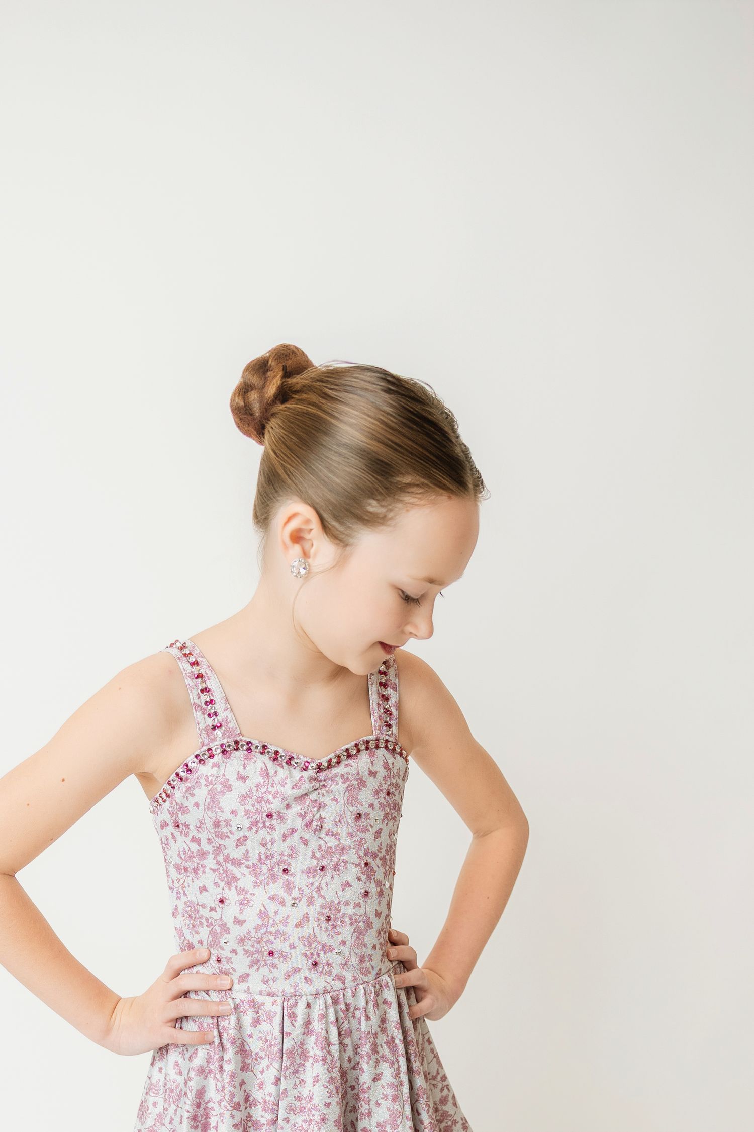 Young girl in floral sundress with hair in bun looking down gracefully against white background.