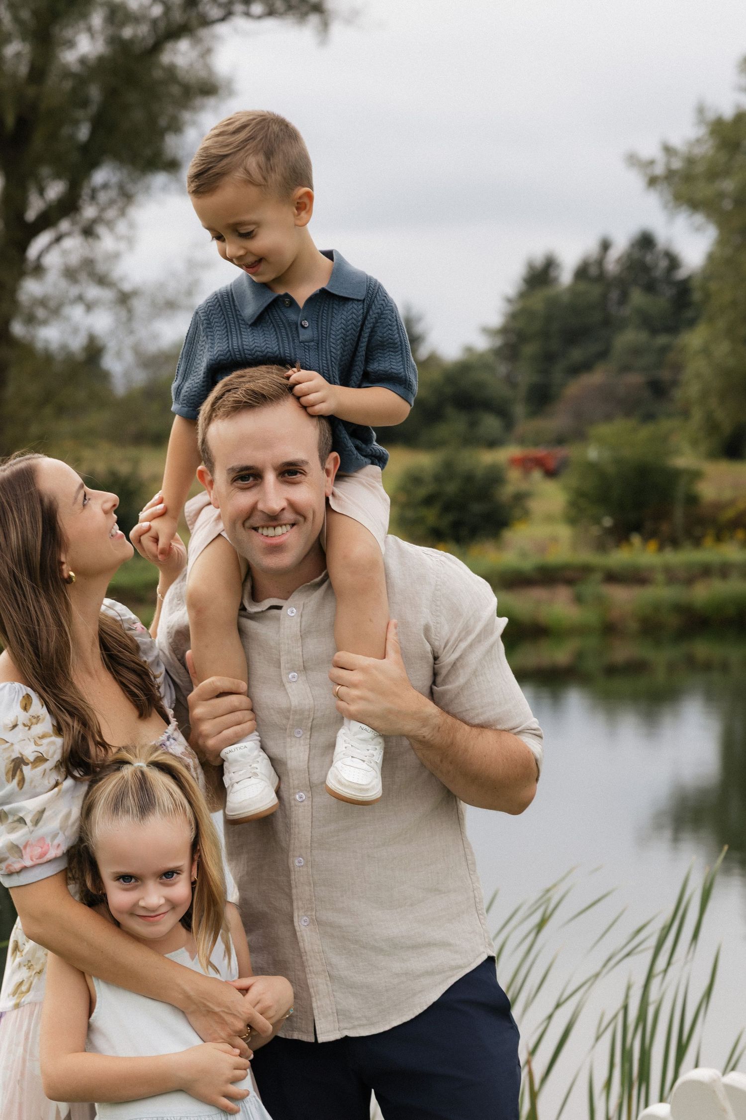 Happy family of four posing outdoors near a pond, father carrying son on shoulders with mother and daughter smiling.