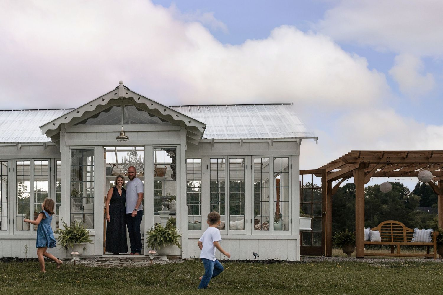 Family posing outside a white greenhouse with children playing on green lawn under cloudy sky.