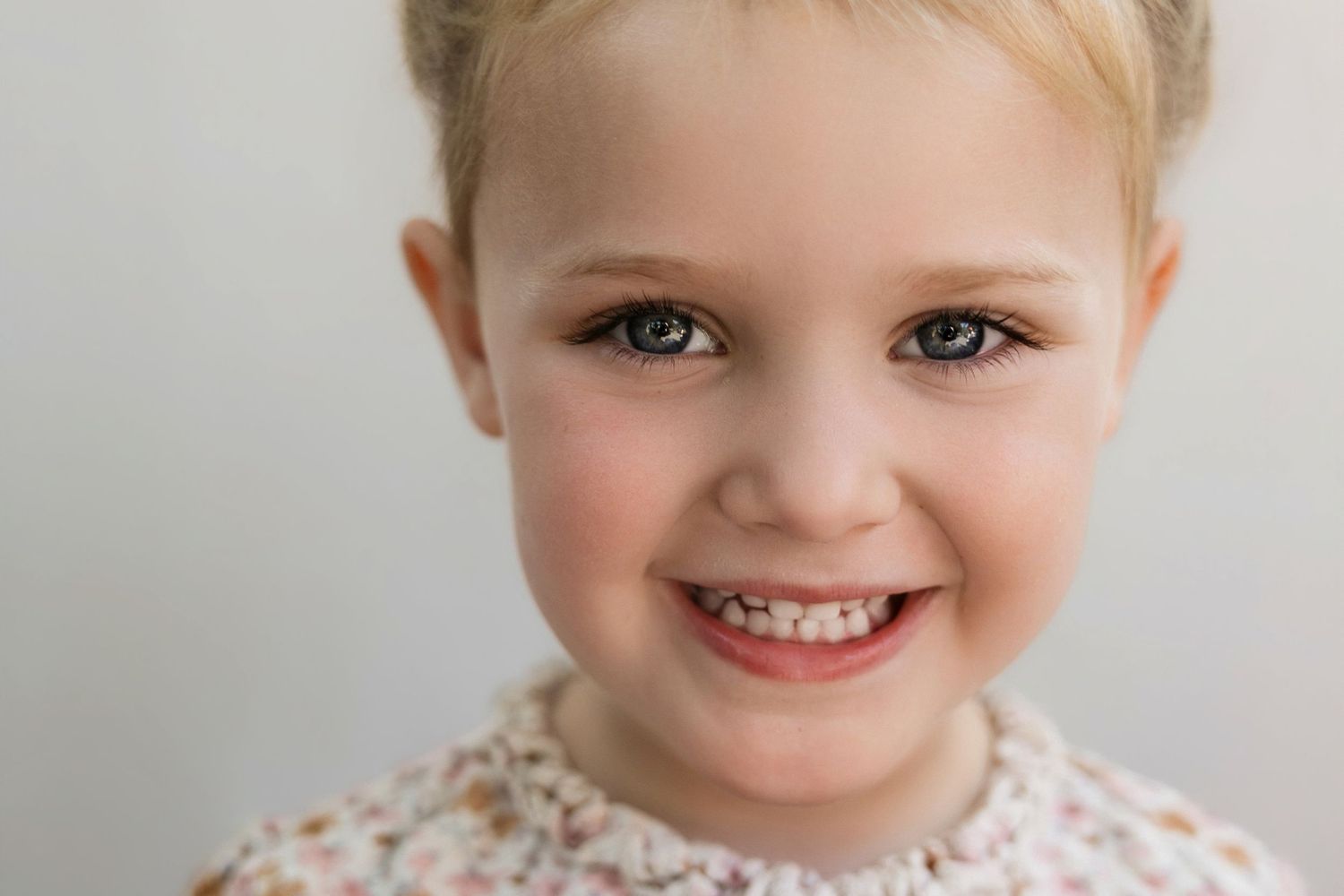 Smiling blonde toddler girl with blue eyes wearing a floral shirt, close-up portrait photo.