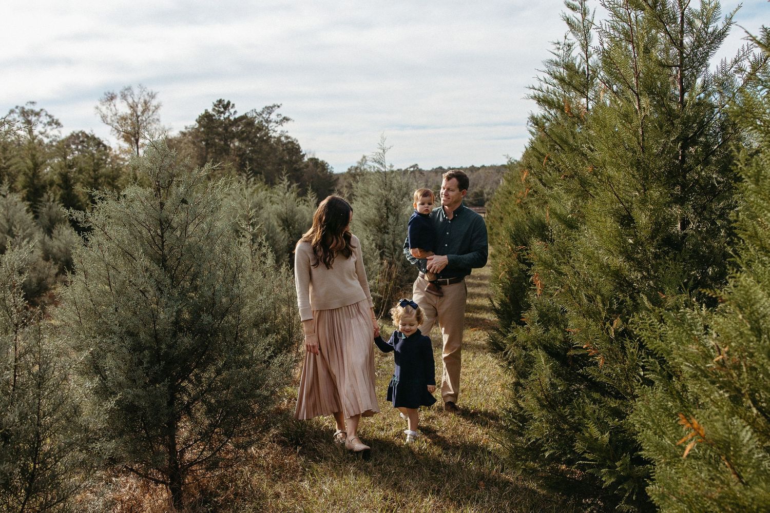 Family of four walking through rows of Christmas trees at a festive tree farm on a cloudy day.