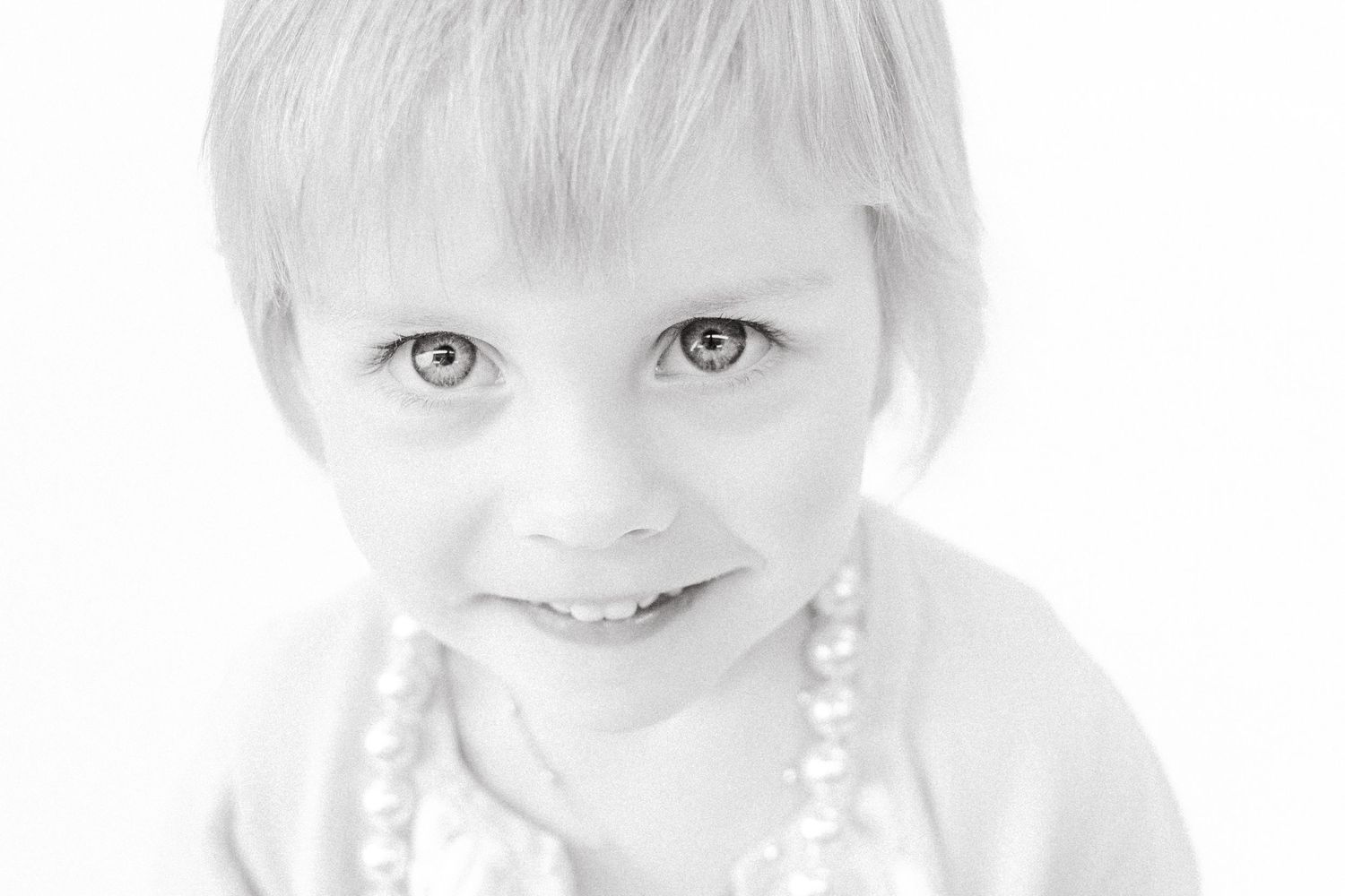 Black and white close-up portrait of a young child wearing a pearl necklace smiling at the camera.