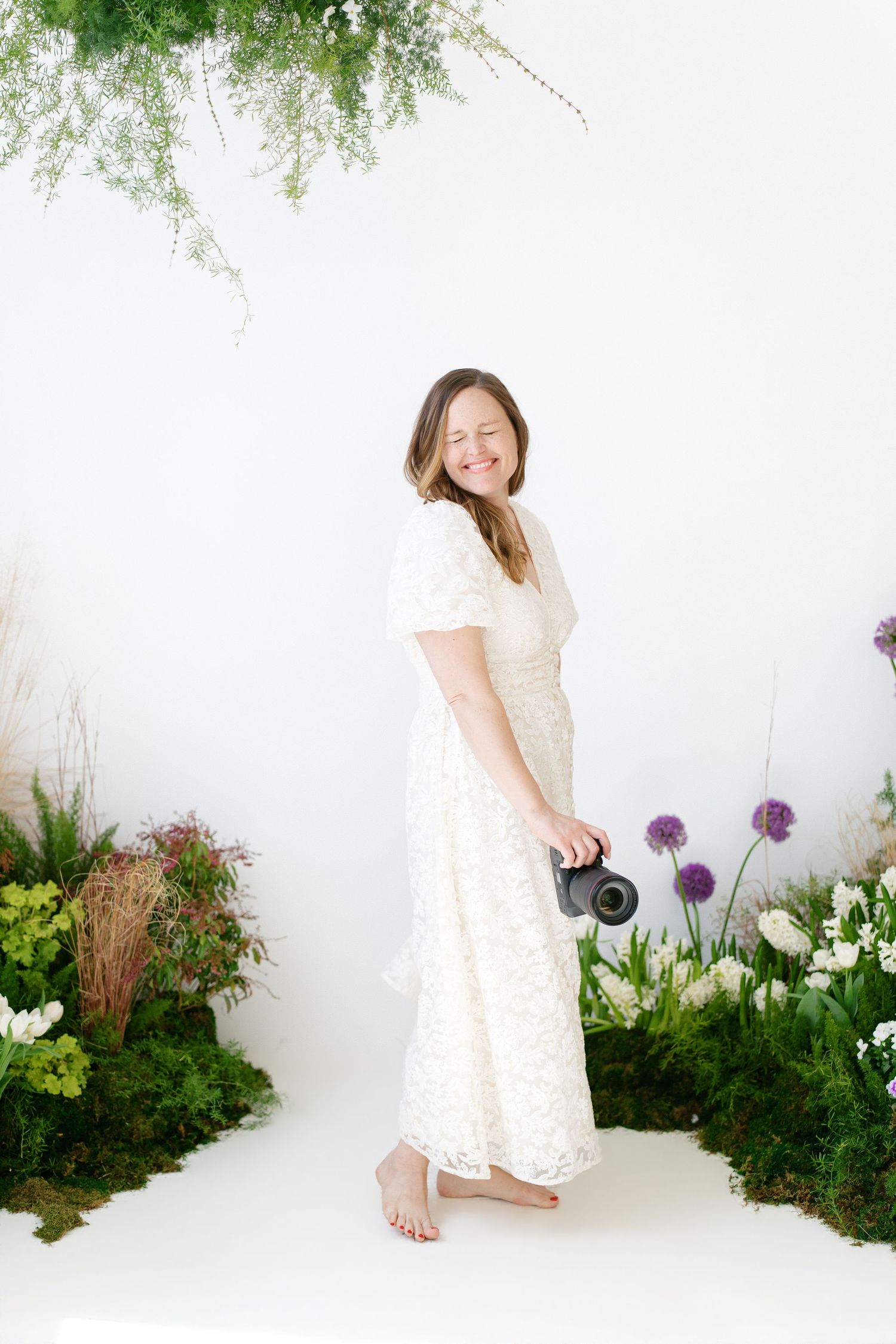Smiling Hilary Mauro in white lace dress holding camera surrounded by flowers.