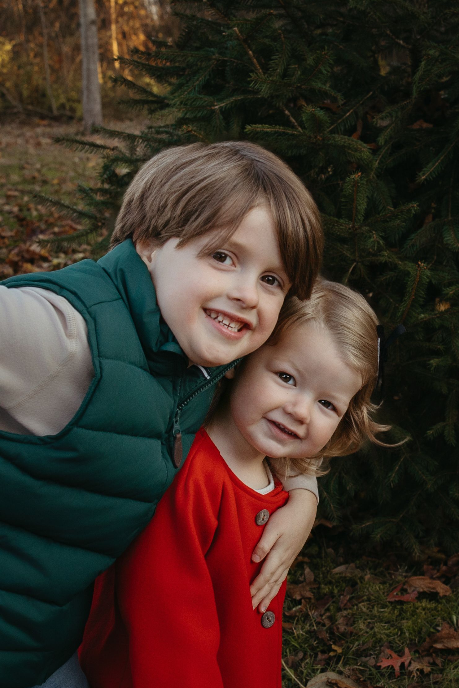 Two smiling young siblings hugging in front of an evergreen tree during an outdoor fall family photo session.