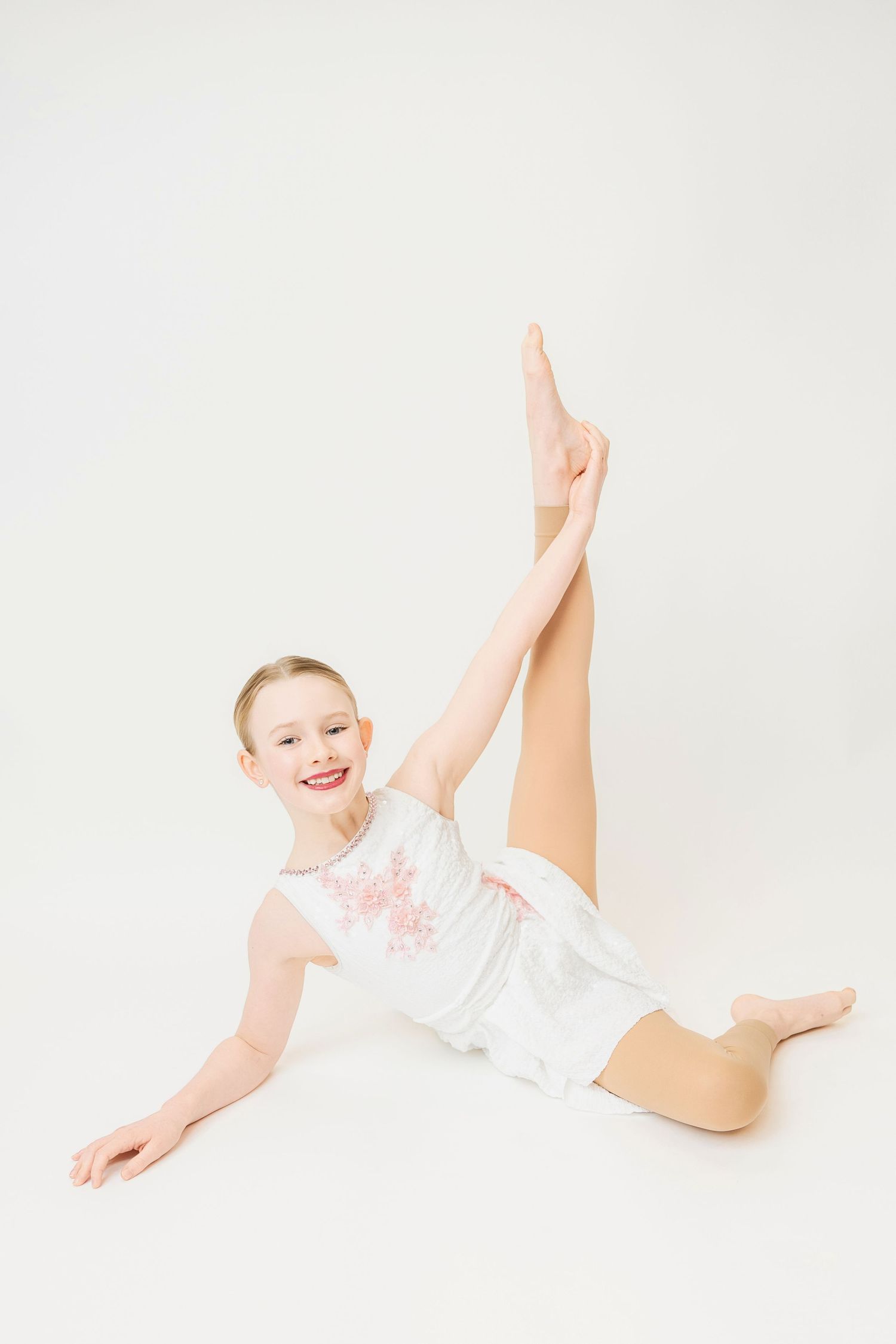Young girl gymnast in white leotard performing a split stretch with leg raised high in a studio.