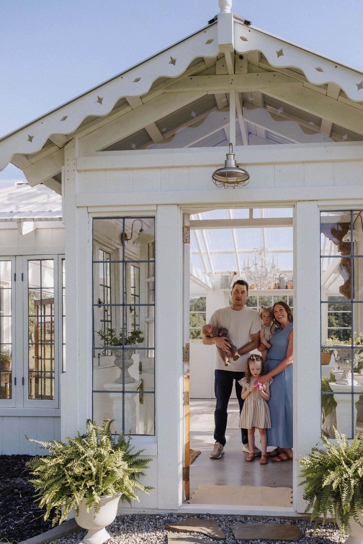 Family of four posing in doorway of charming white glass greenhouse with ferns and blue sky backdrop.