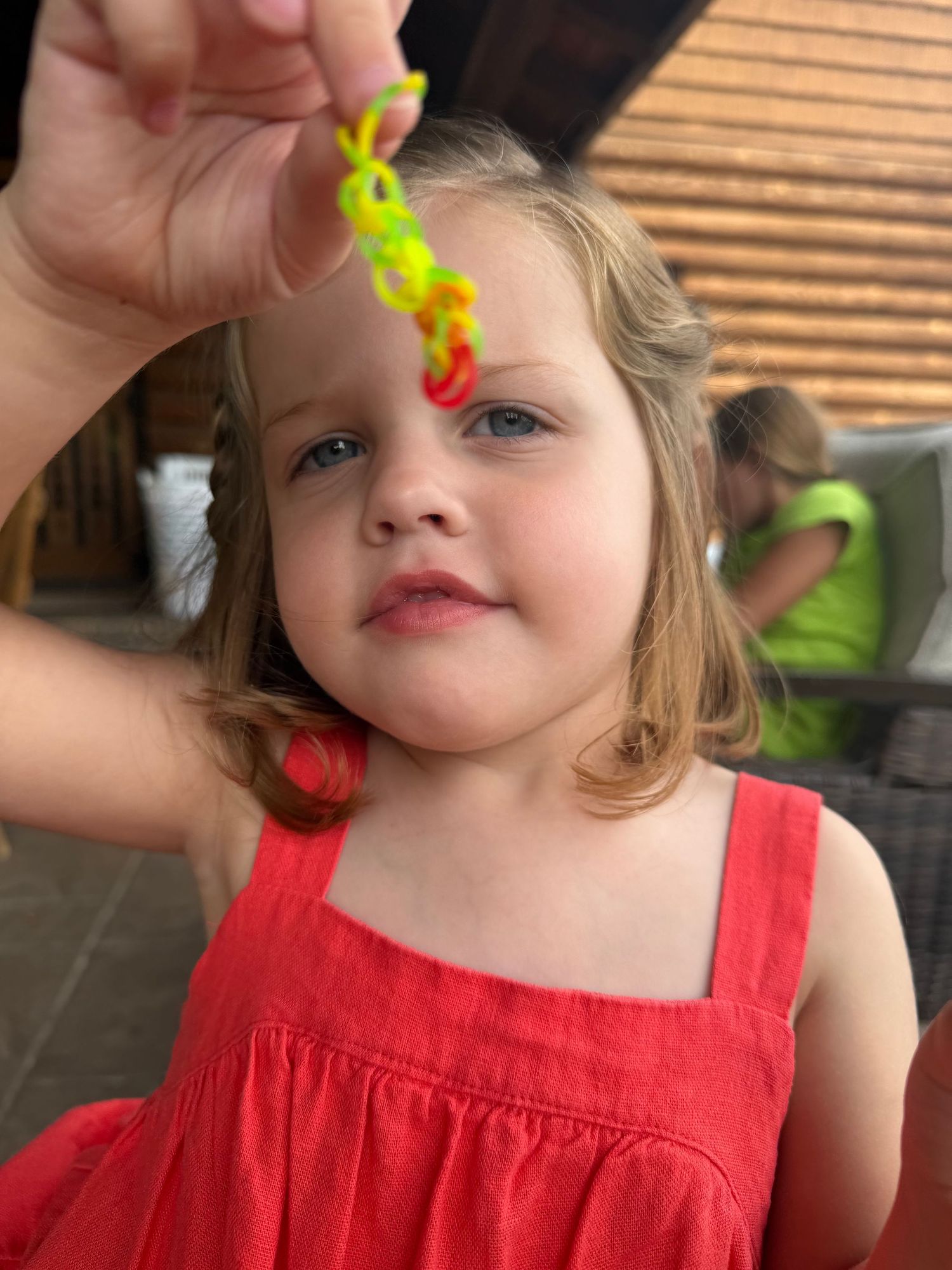 A smiling toddler girl in a red dress holds up a colorful yellow and red rubber band loom bracelet outdoors.