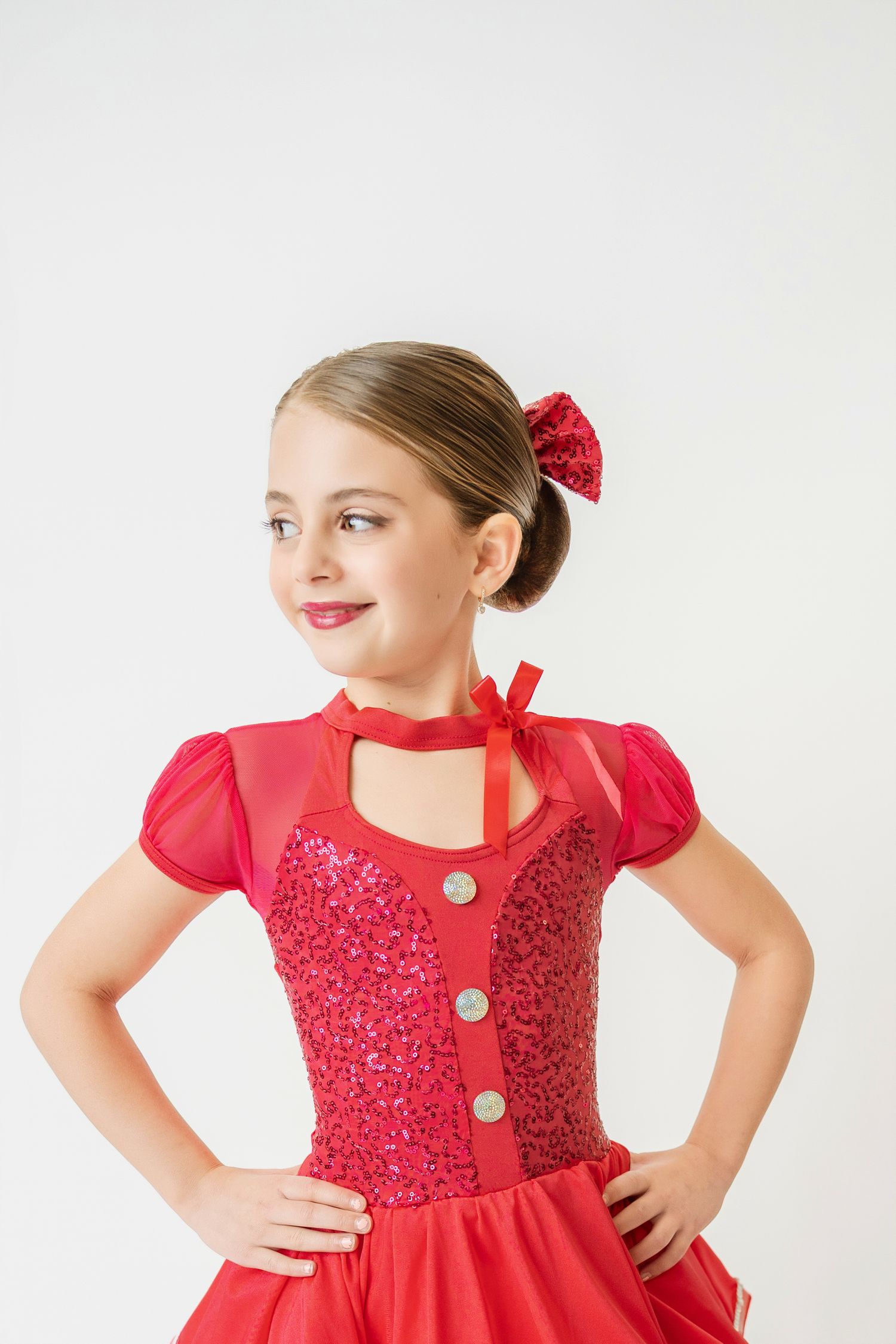 Young girl in red sequin dance costume with puff sleeves smiling confidently with hands on hips.