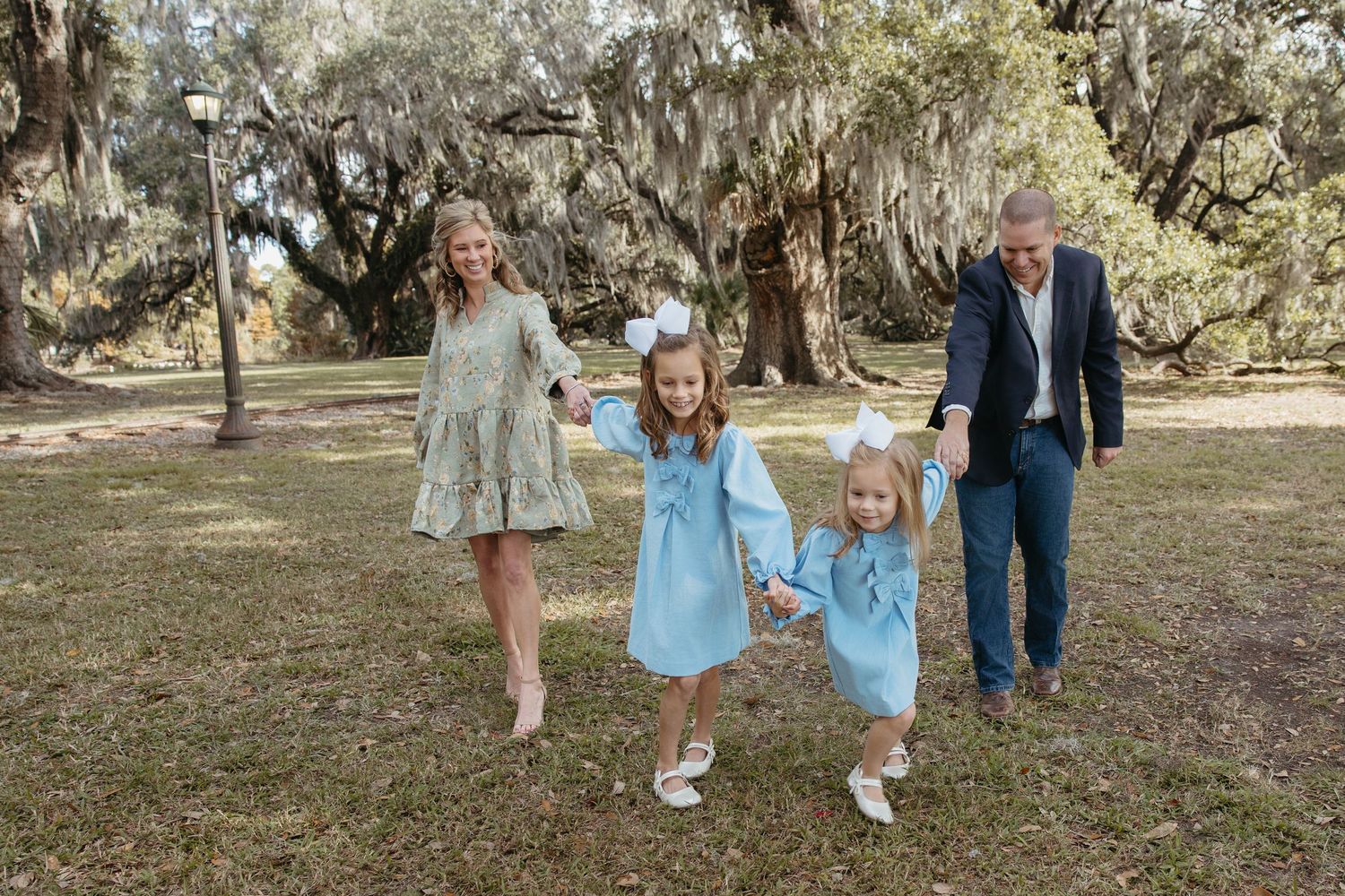 Family of four holding hands and walking under Spanish moss-draped trees in a park.