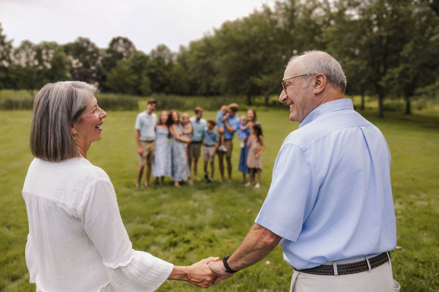 Elderly couple holding hands and smiling in a green park with family gathered in the background.