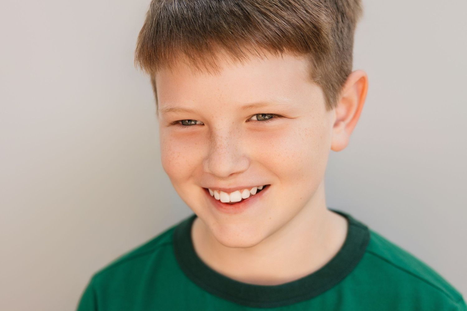 Smiling young boy with short brown hair wearing a green t-shirt against a light gray background.
