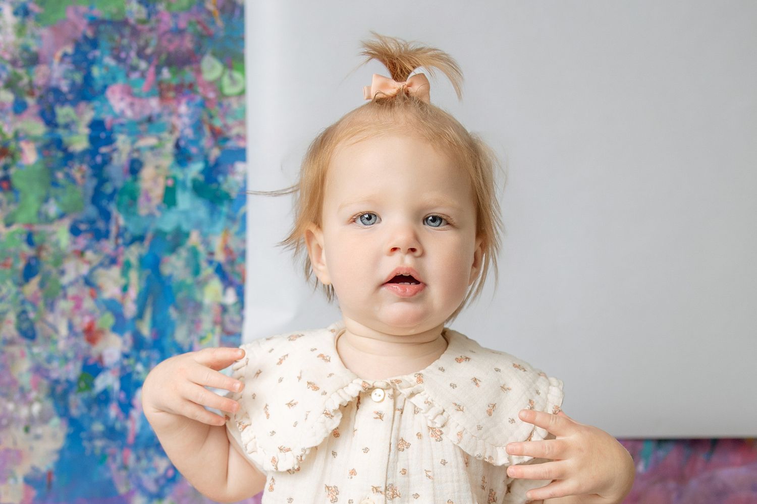 Adorable toddler girl with a tiny hair bow in a floral dress standing before colorful abstract artwork.