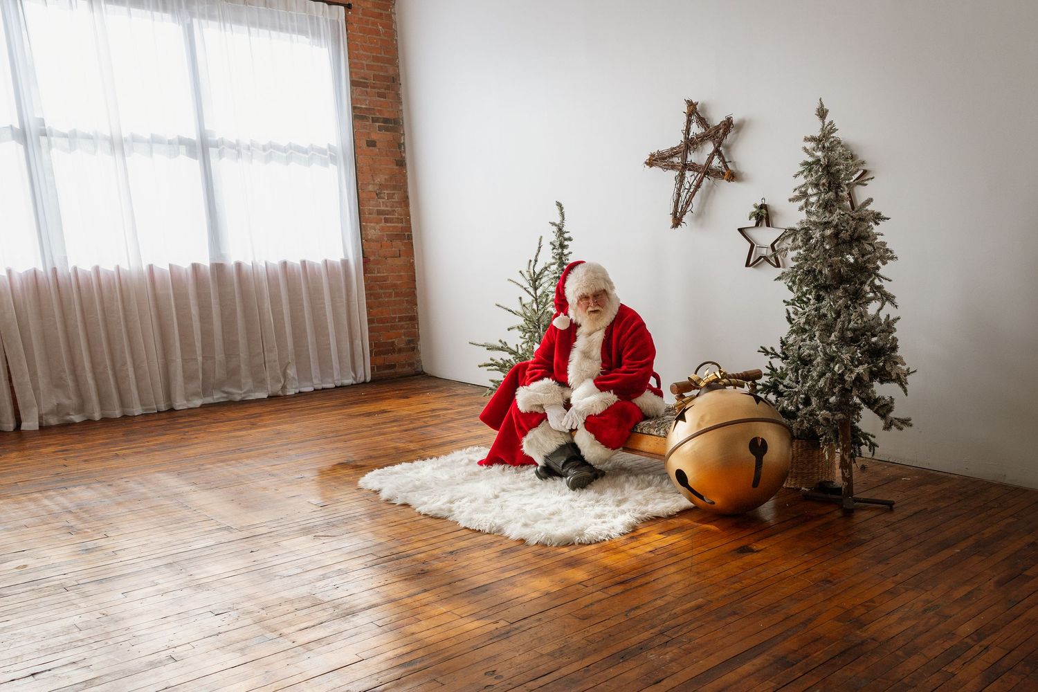 Santa Claus kneels beside a large golden bell ornament near a frosted Christmas tree indoors.