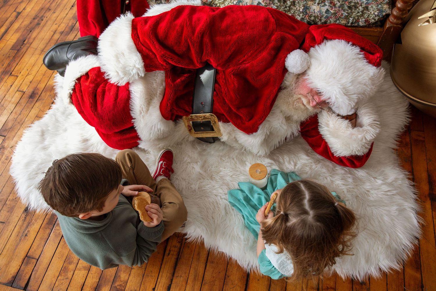 Overhead view of two children sharing cookies with Santa Claus on a white fur rug at Christmas.