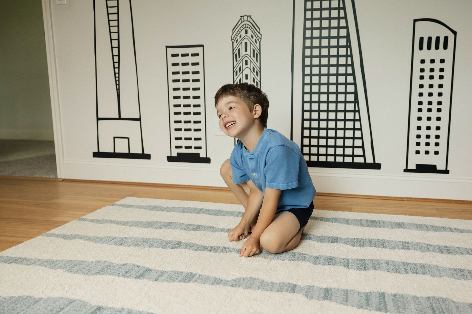 Happy young boy kneeling on a striped rug in front of a city skyline mural indoors.