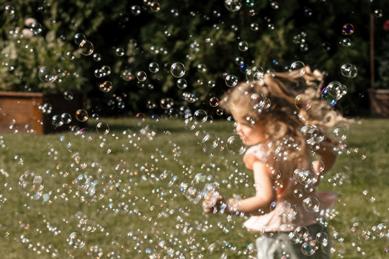 A young girl with curly blonde hair joyfully runs through a yard filled with floating soap bubbles on a sunny day.