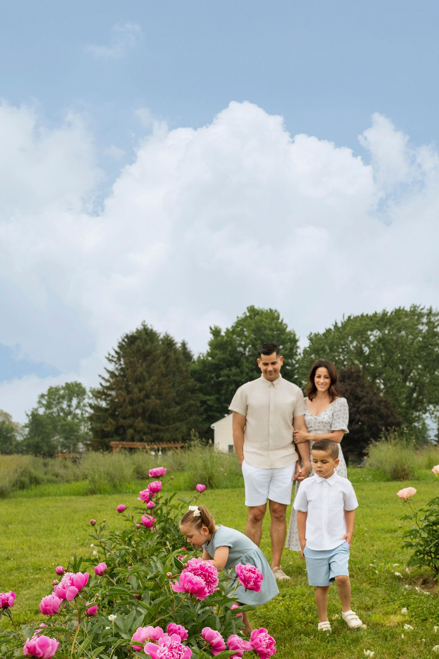 Family of four enjoying a sunny day in a garden filled with blooming pink peonies and green trees.