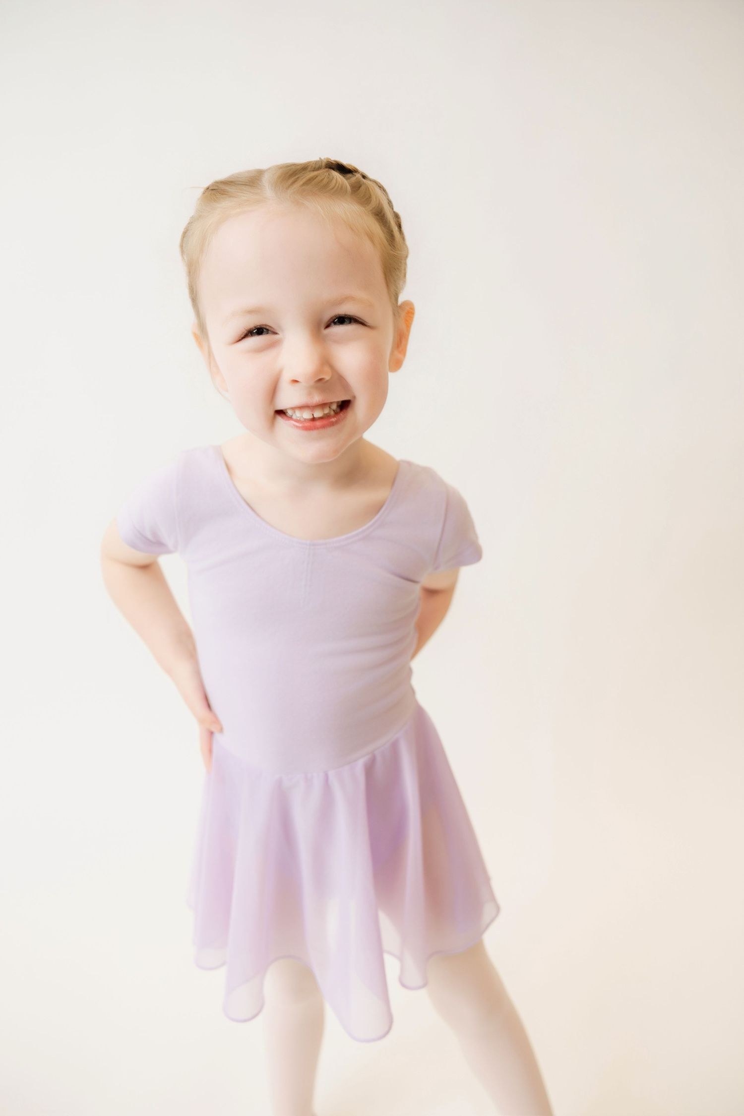 Smiling young girl in lavender ballet leotard and skirt posing with hands on hips in dance studio.