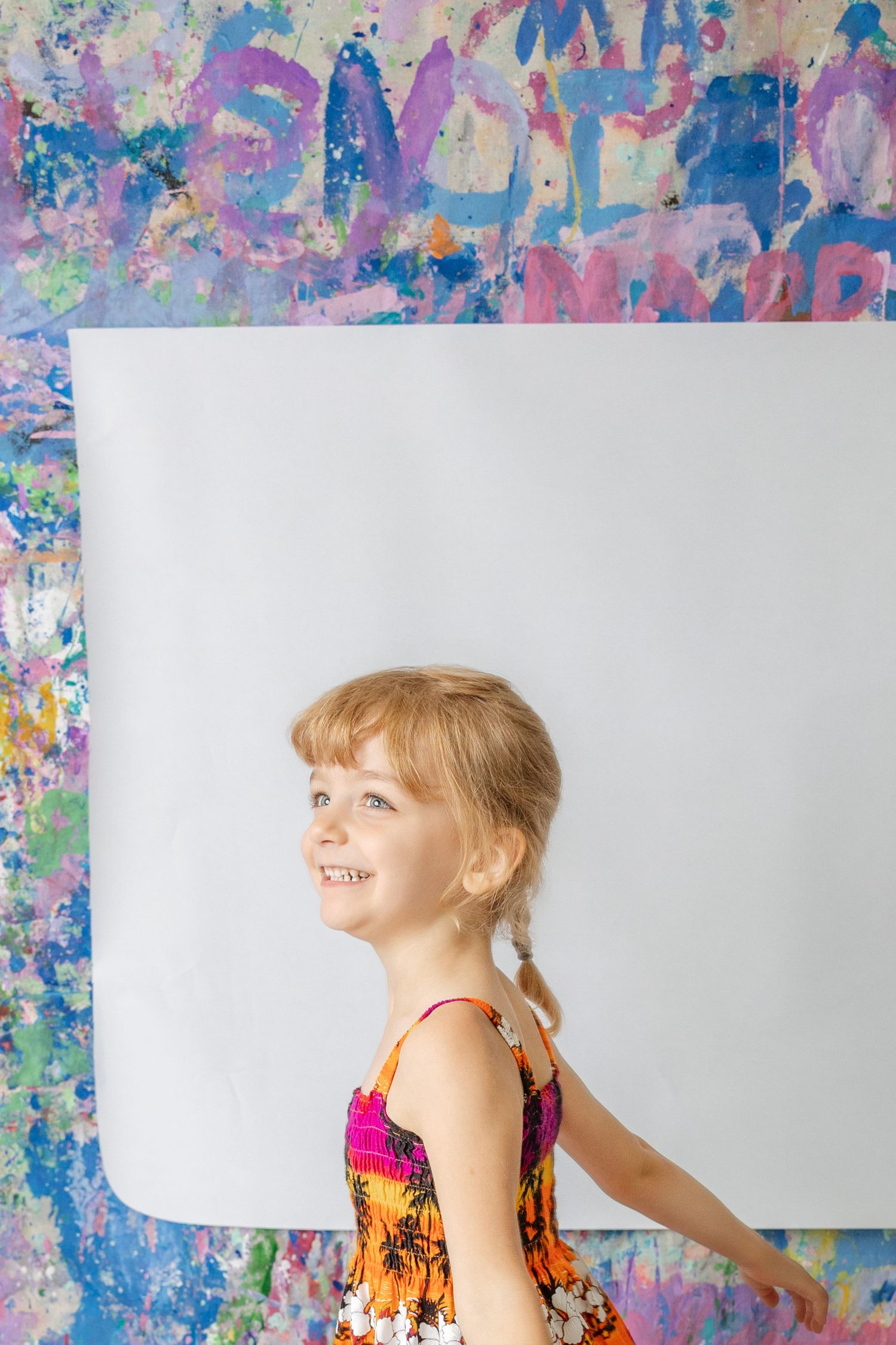 Smiling young girl in floral dress standing in front of colorful painted wall with blank white canvas.