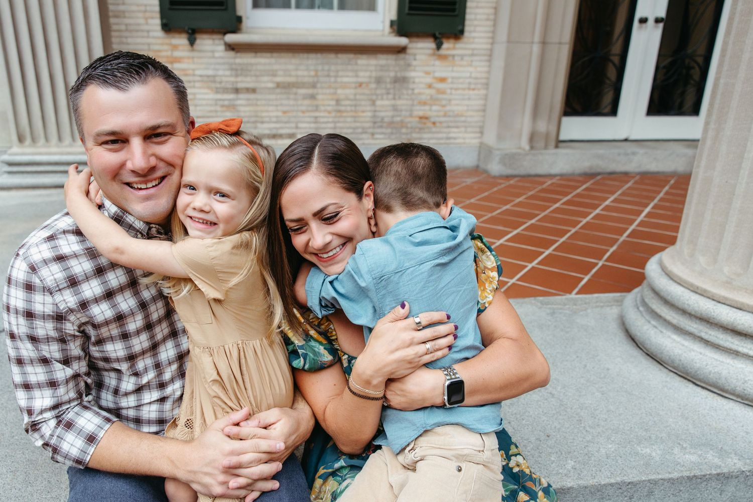 Happy family of four sharing hugs and laughter on steps of a classic brick building.