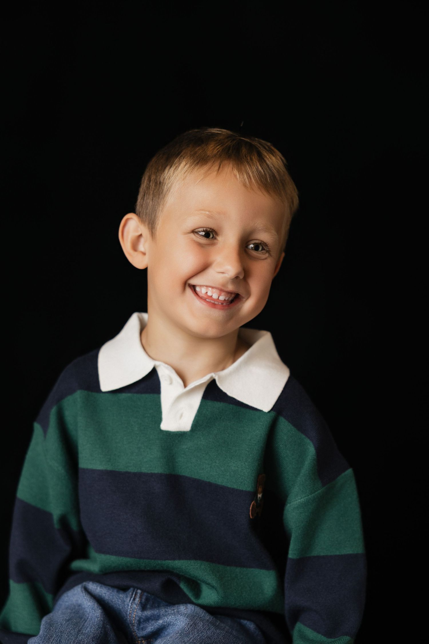 Smiling young boy in a green and navy striped polo shirt poses against a black background.
